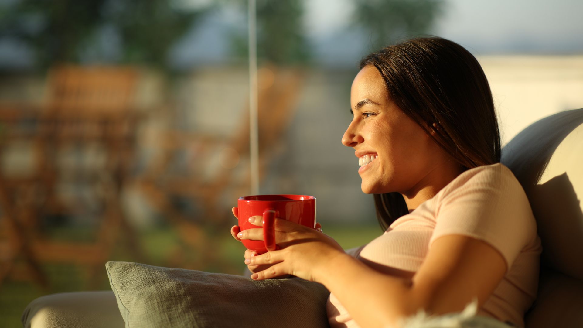 A woman relaxes on a couch with a warm red mug in hand, smiling as she looks outside, capturing a moment of peace and security—perhaps reflecting the reassurance that comes from having a Durable Power of Attorney in place for future decision-making.