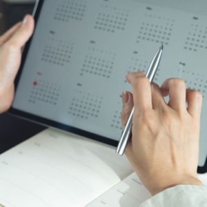 A person holds a tablet displaying a digital calendar while pointing at a date with a pen, possibly scheduling an important legal task such as setting up a Durable Power of Attorney or managing estate planning deadlines